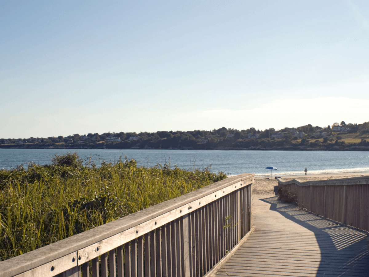 A wooden boardwalk leads to a sandy beach with tall grasses on the left and calm water in the background. The sky is clear and sunny.