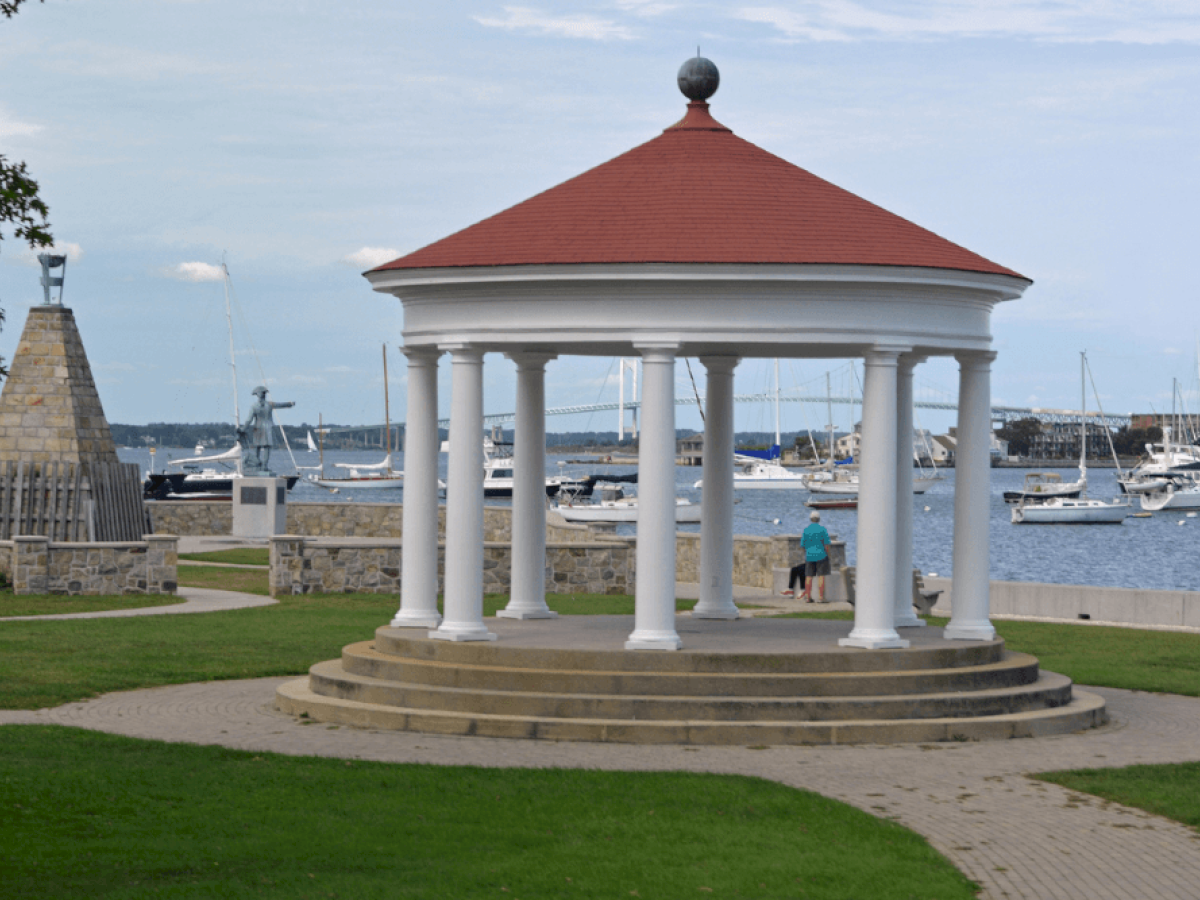 A gazebo with a red roof overlooks a marina with sailboats. A stone monument is on the left, and two people stand inside the gazebo.