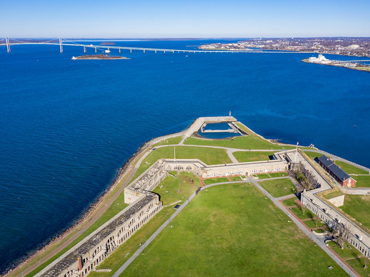 Aerial view of Fort Adams with a wide expanse of grass, surrounded by water and a bridge in the background, under a clear blue sky.