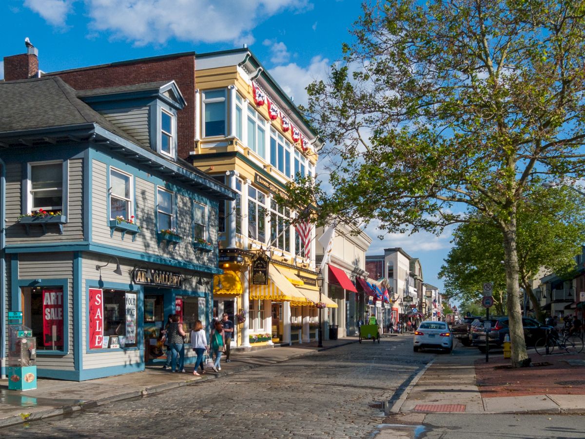 A charming street scene with colorful shops, people walking, trees, and cars parked, under a blue sky with a few clouds.