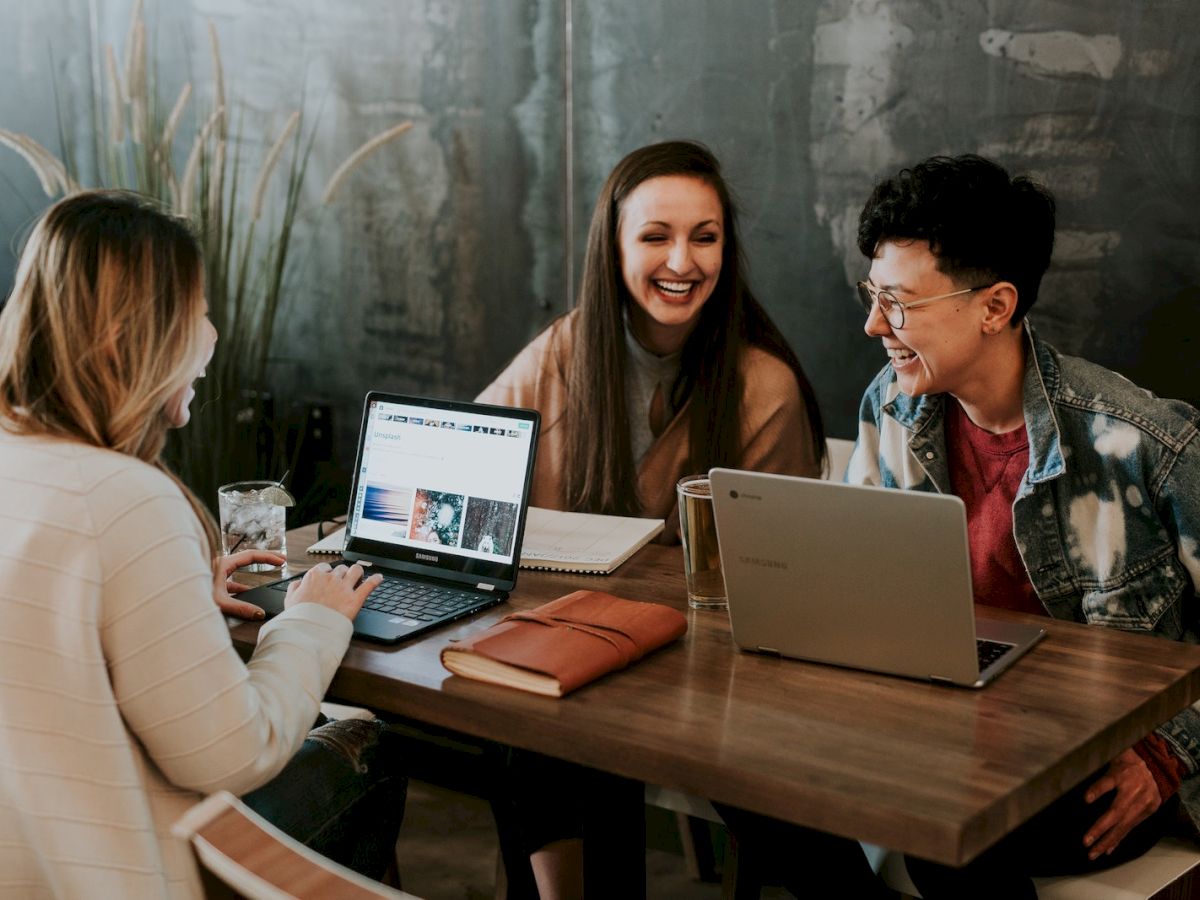 Three people are sitting at a table with laptops, notebooks, and drinks, engaged in conversation and smiling together.