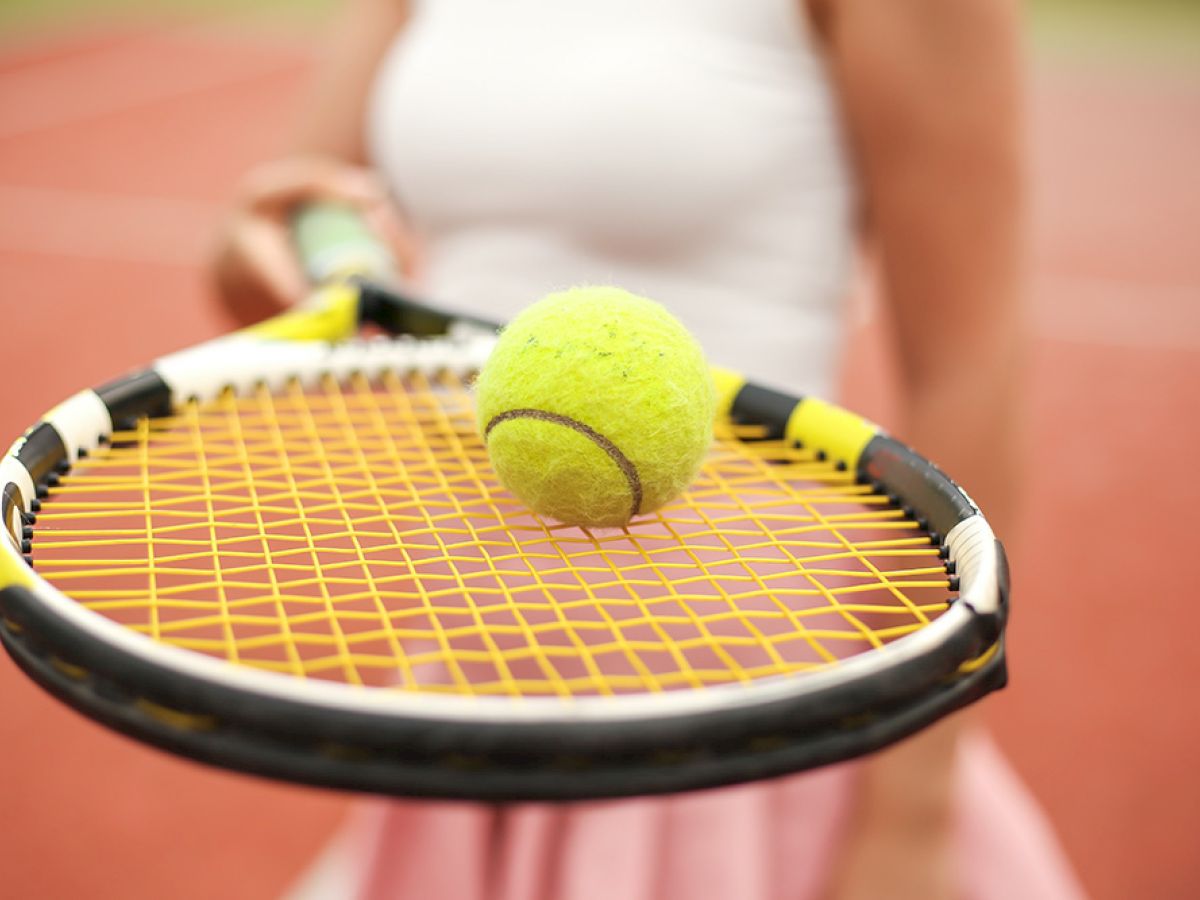 A person holding a tennis racket with a tennis ball balancing on it on a court.