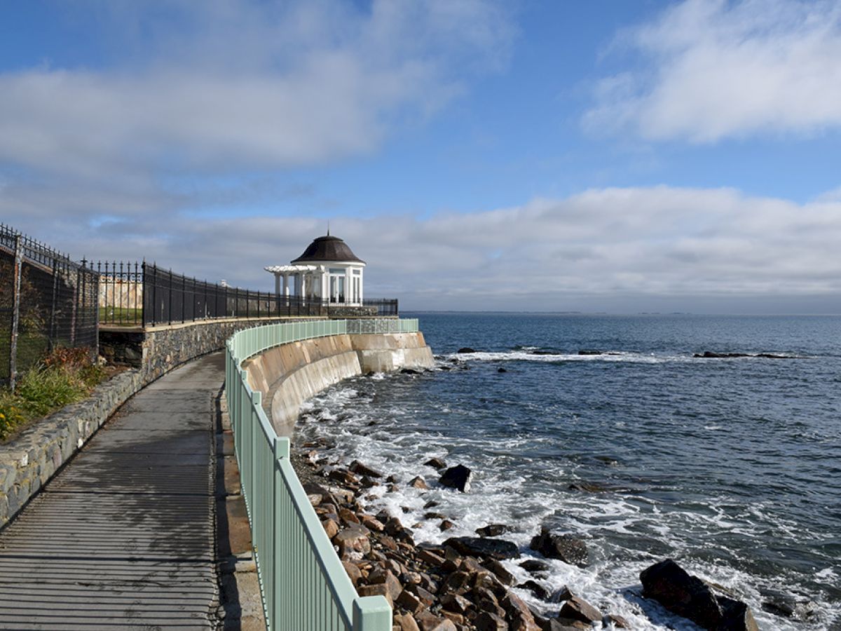 A coastal path with a railing runs alongside the ocean, leading to a small gazebo. Waves crash against the rocky shore under a partly cloudy sky.