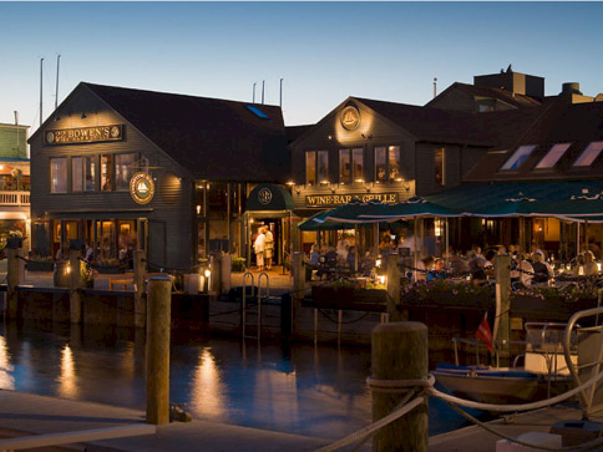 A waterfront dining area at dusk, with people seated outdoors. The scene reflects a cozy, lively atmosphere by the water's edge.