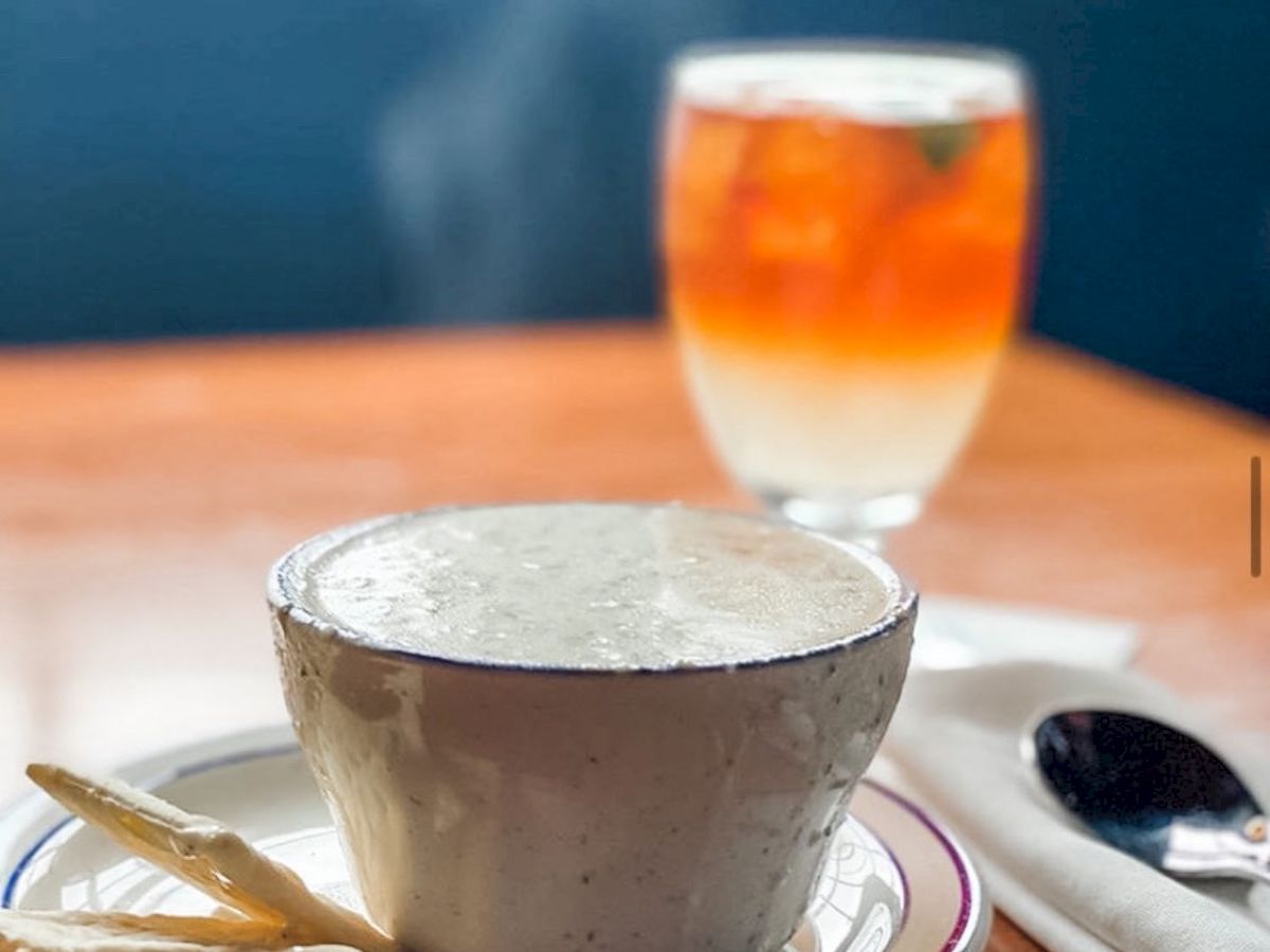 A steaming bowl of soup with crackers on the side, and a glass of iced tea in the background on a wooden table.