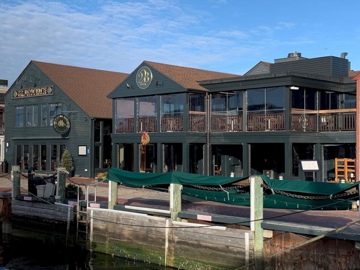 The image shows a waterfront building with large windows and green accents, featuring a dock in the foreground and a sign on the facade.