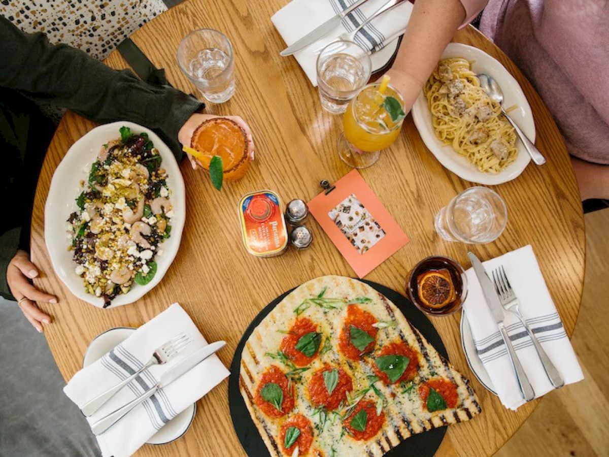 A table with a salad, pasta, flatbread, drinks, and cutlery, with two people sitting.