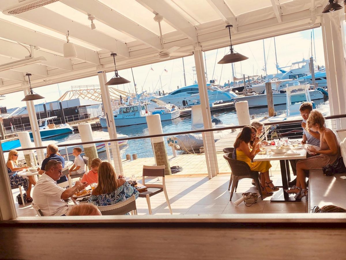 People are dining at a waterfront restaurant with boats docked outside under a covered patio.