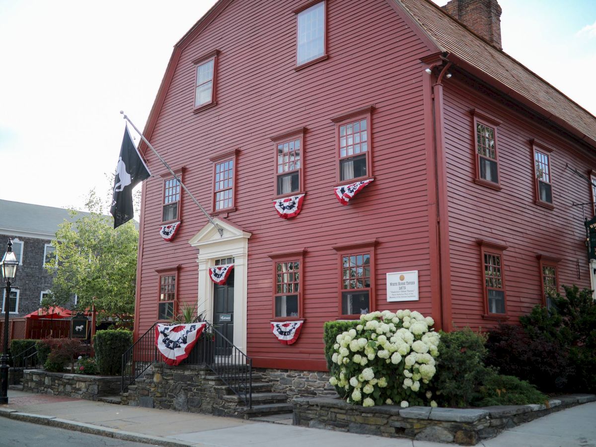 A historical red building adorned with patriotic bunting, a black flag, and a sign. Lush greenery enhances its charming facade.