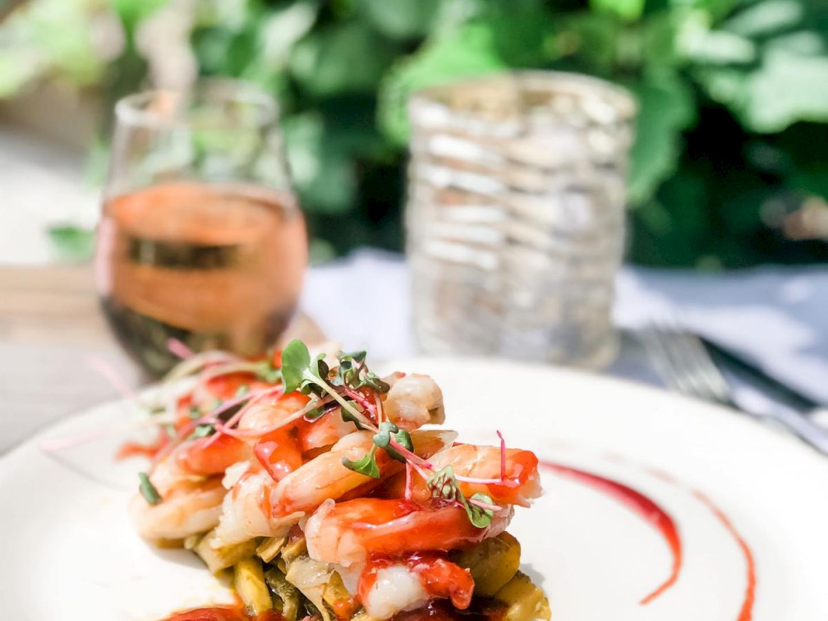 A gourmet dish with layered vegetables and shrimp, garnished with microgreens, served on a plate with sauce, next to a glass of rosé wine.