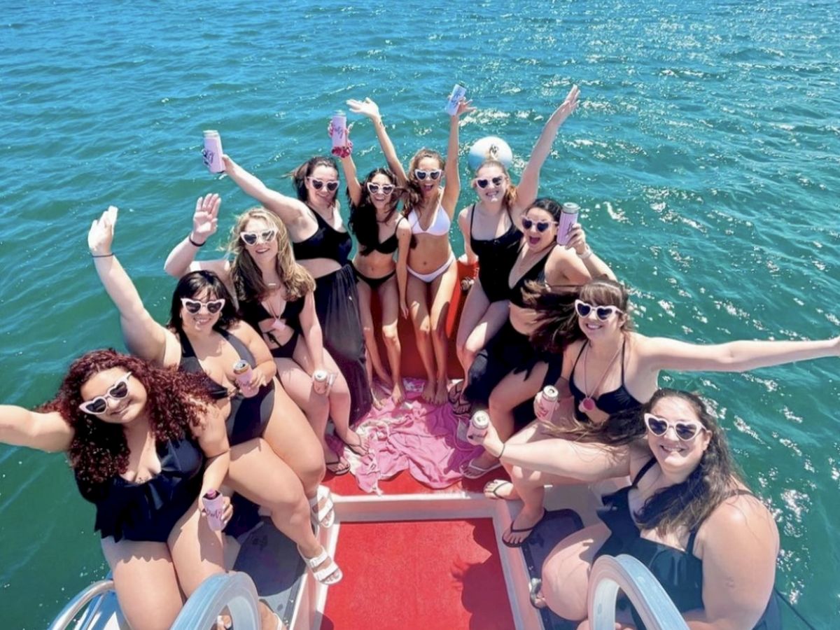 A group of people in swimsuits are on a boat, smiling and holding drinks, with a body of water and another boat in the background.