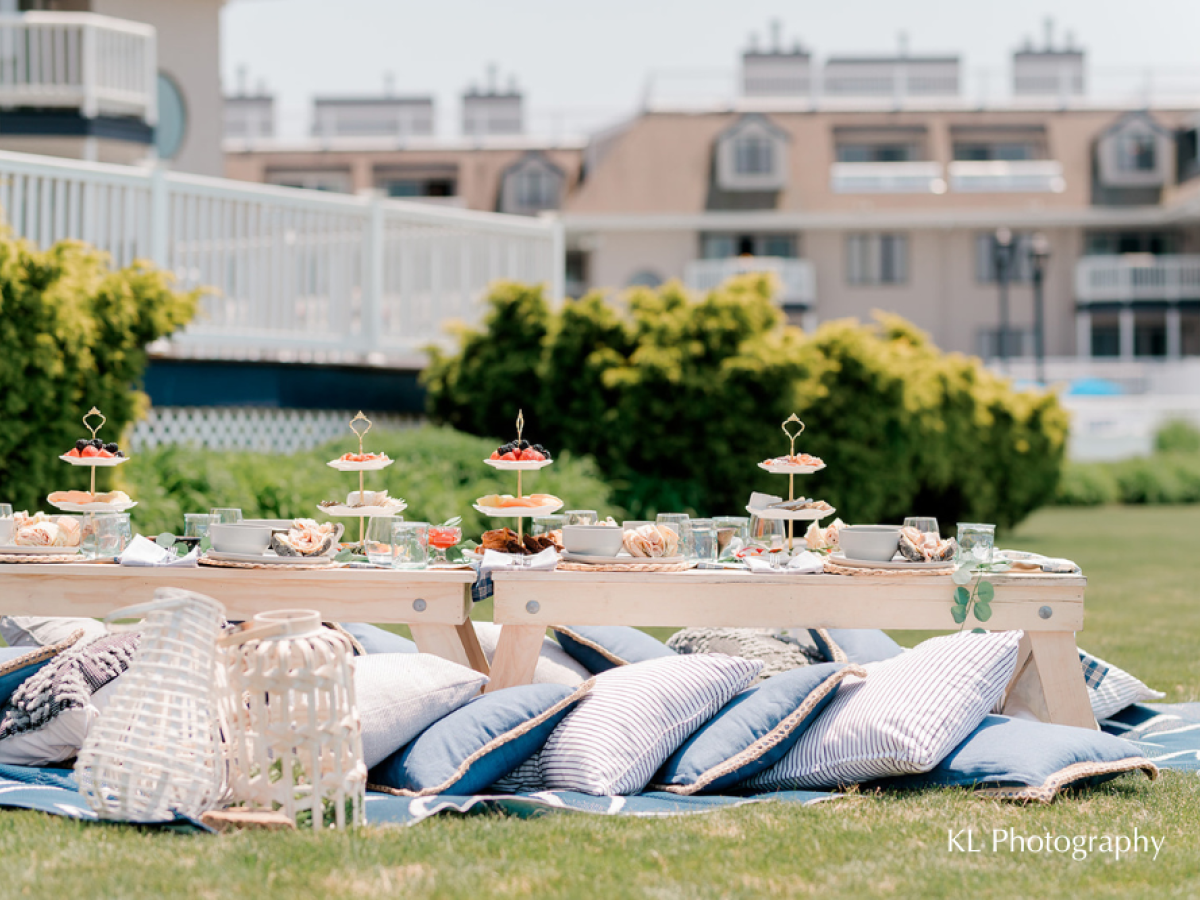 An outdoor picnic setup with a low table, cushions, tiered trays of snacks, and lanterns, set on a lawn near a building.