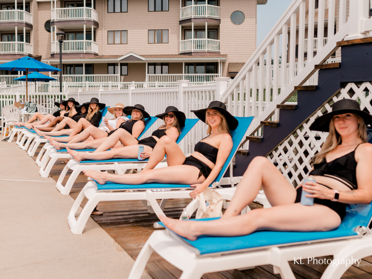 A group of women in black swimsuits and hats lounge on chairs by a pool, with a building in the background.