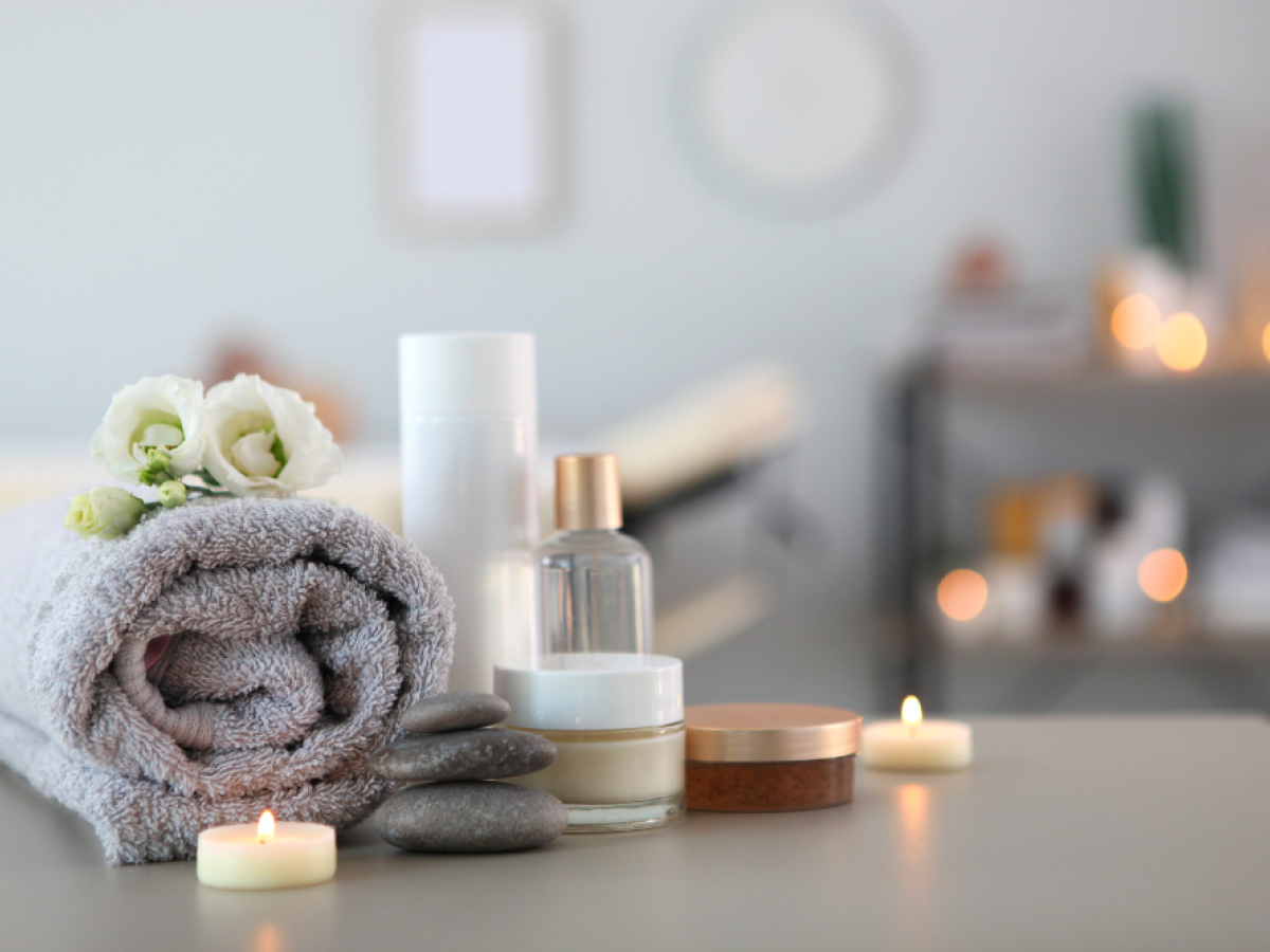 A spa setting with a rolled towel, candles, stones, and beauty products on a table, creating a relaxing atmosphere.