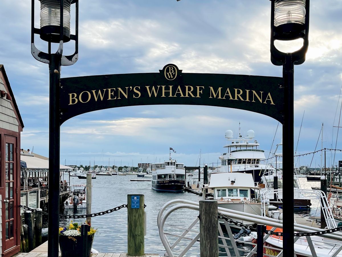 The image shows the entrance to Bowen's Wharf Marina with boats docked and a seagull flying above, under cloudy skies.