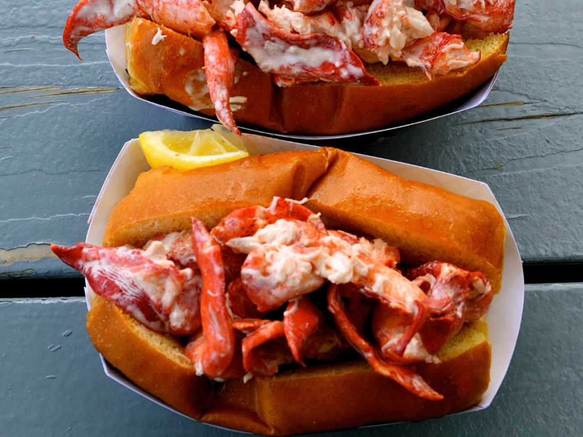 Lobster rolls filled with lobster meat on a toasted bun, with a lemon wedge, served in paper trays on a blue wooden table.