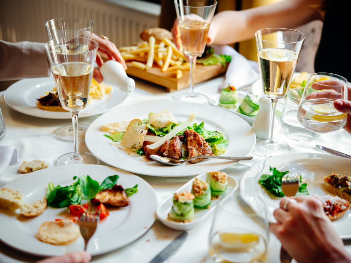 A dining table with various dishes, including steak and vegetables, surrounded by glasses of champagne, and people enjoying the meal.