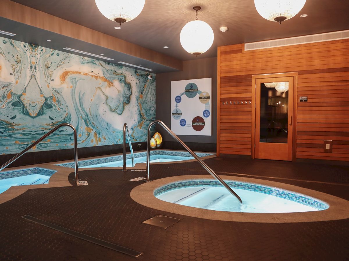 The image shows a spa area with hot tubs, a sauna, and a striking wall mural, illuminated by round ceiling lights.