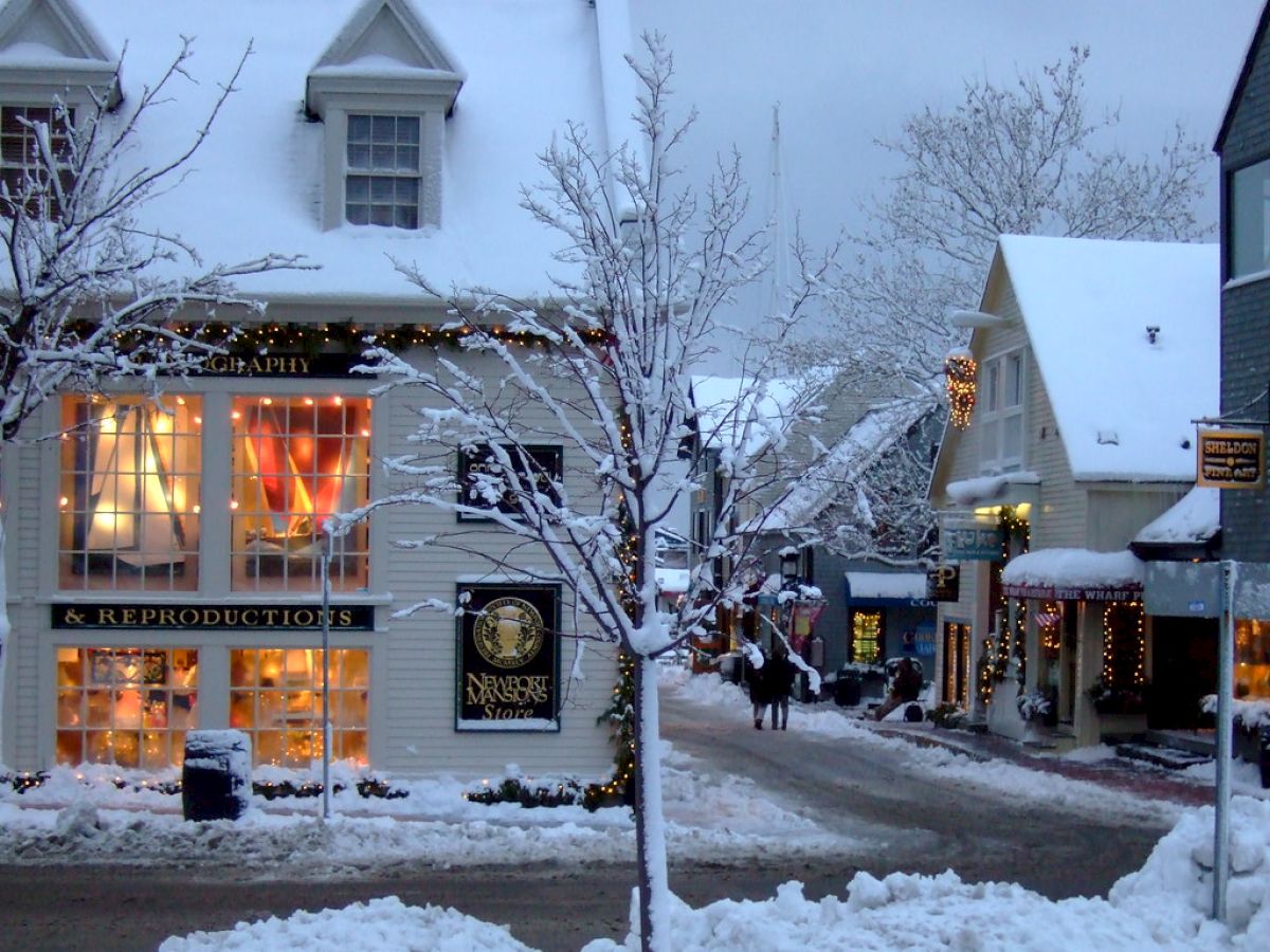 A snowy street scene with snow-covered buildings, lighted windows, and bare trees, creating a cozy winter atmosphere.