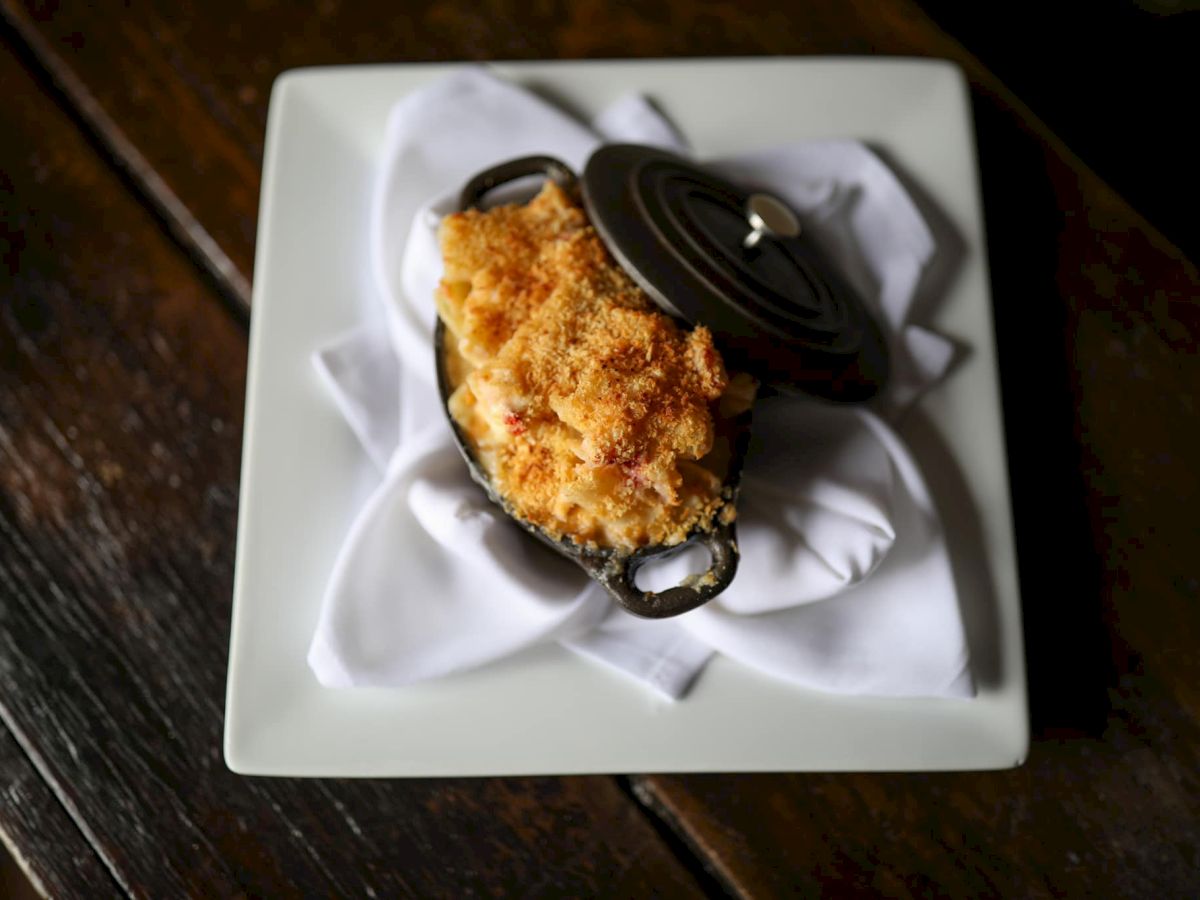 A small dish of baked macaroni and cheese with a breadcrumb topping, served in a mini cast iron pot on a white square plate with a napkin.