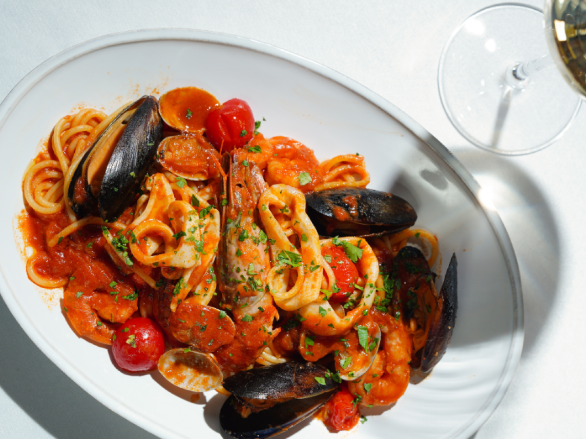 A plate of seafood pasta with mussels, shrimp, spaghetti, and cherry tomatoes in tomato sauce, garnished with herbs. A glass of white wine nearby.