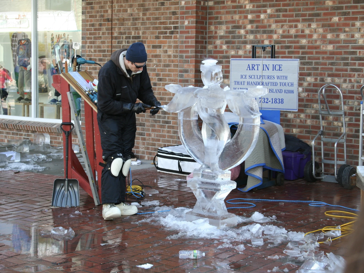 An artist is carving an ice sculpture outside, with tools scattered around. A sign behind them reads 