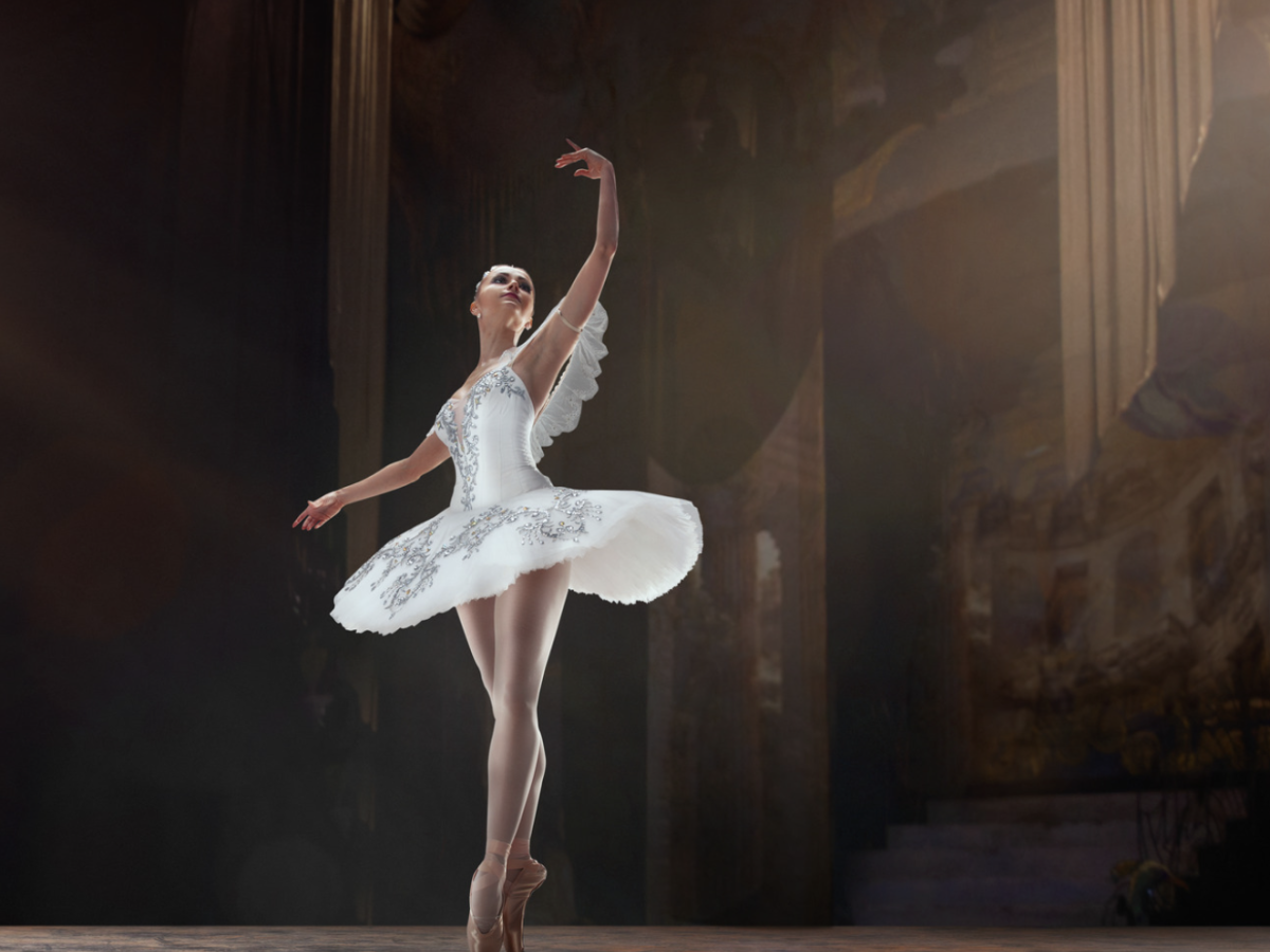 A ballerina in a white tutu performs on stage with dramatic lighting and a painted backdrop, striking an elegant pose.