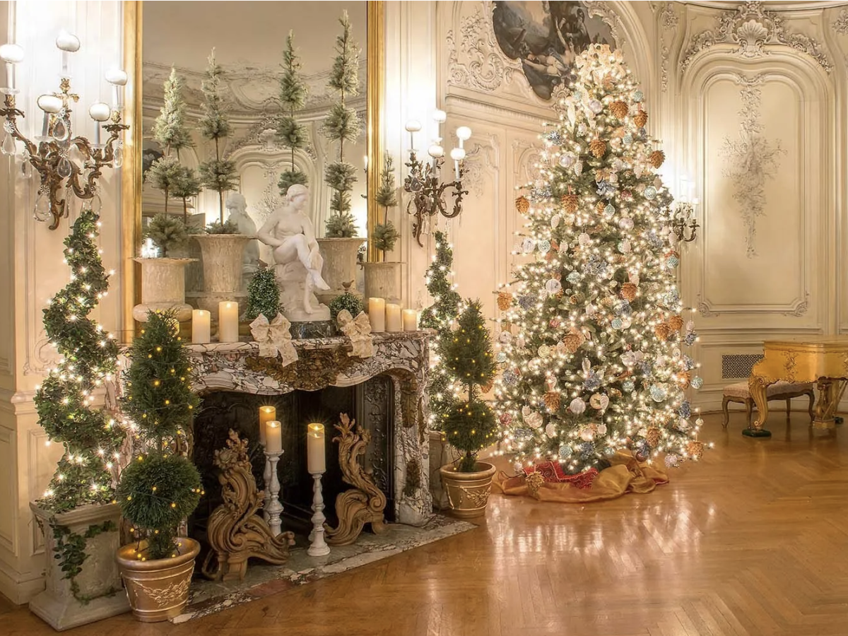 A lavishly decorated room with a lit Christmas tree, ornate fireplace, candles, topiary trees, and elegant furnishings completes the scene.