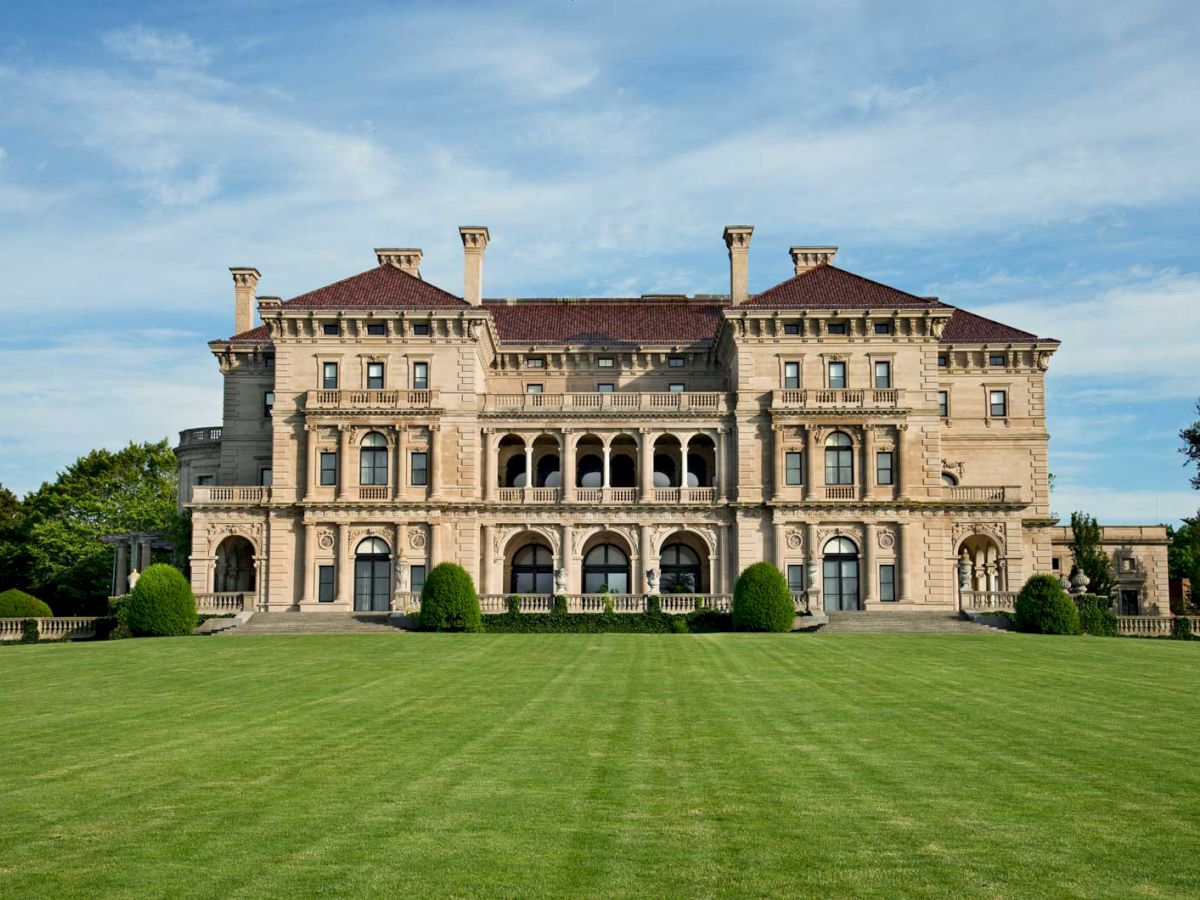 The image shows a large, historic mansion with ornate architecture, expansive lawns, and clear skies in the background.