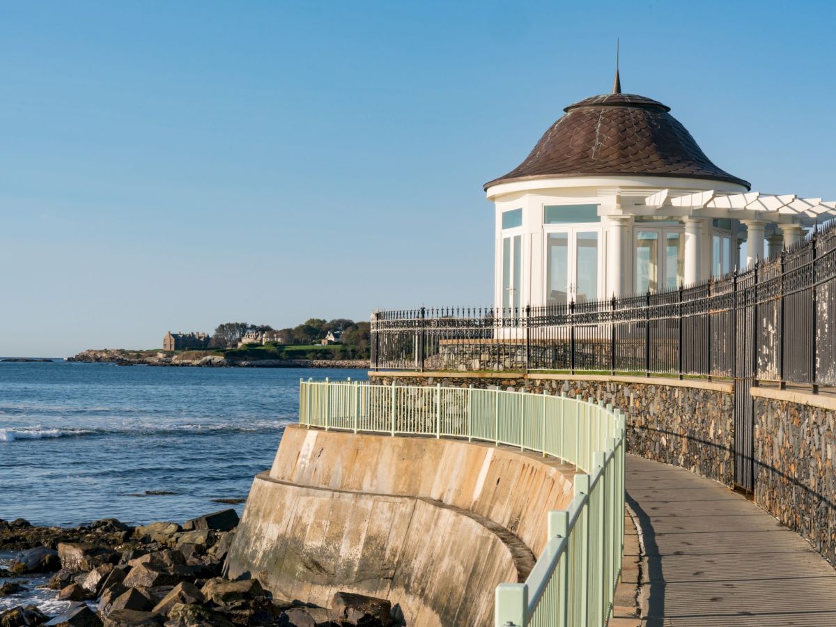 A coastal walkway with a rotunda overlooks the ocean, lined with railings and bordered by rock formations and sunny skies.