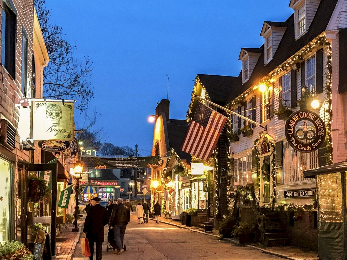 A cozy street scene in the evening with lit storefronts, festive decorations, and people walking under an American flag.