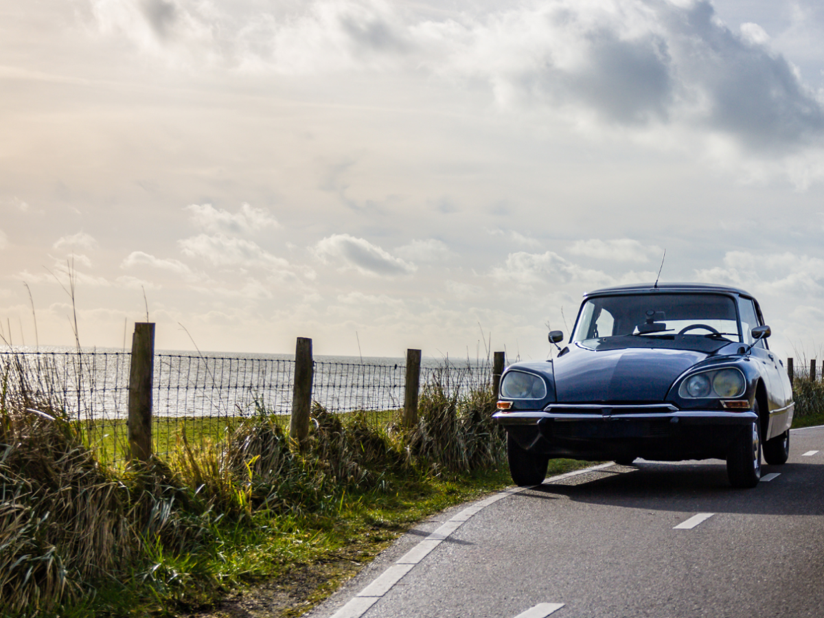 A vintage car drives along a coastal road, flanked by a wooden fence and greenery, with the ocean and cloudy sky in the background.