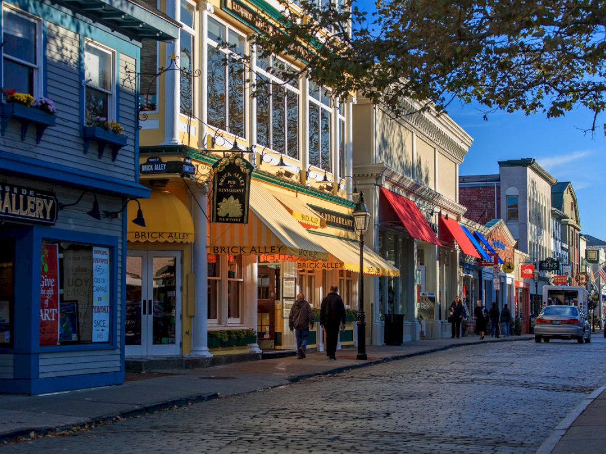 A charming street lined with colorful shops and awnings, people walking, and a vintage car driving along the cobblestone road.