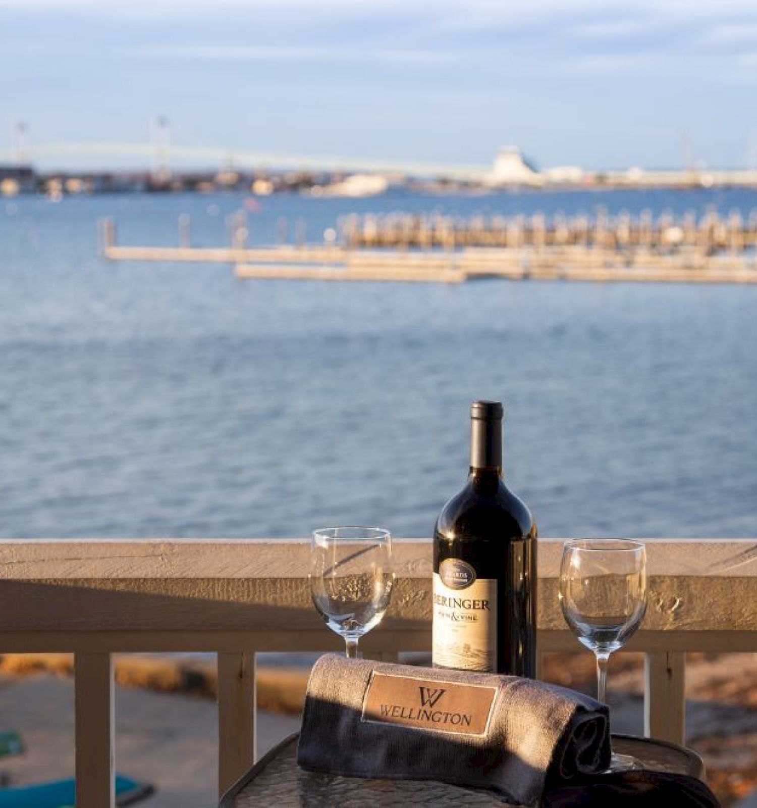 A bottle of wine and two glasses on a table overlooking a waterfront with a pier in the background.