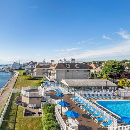 A coastal resort with a pool and blue umbrellas overlooks the water, surrounded by greenery and adjacent buildings, under a clear sky.
