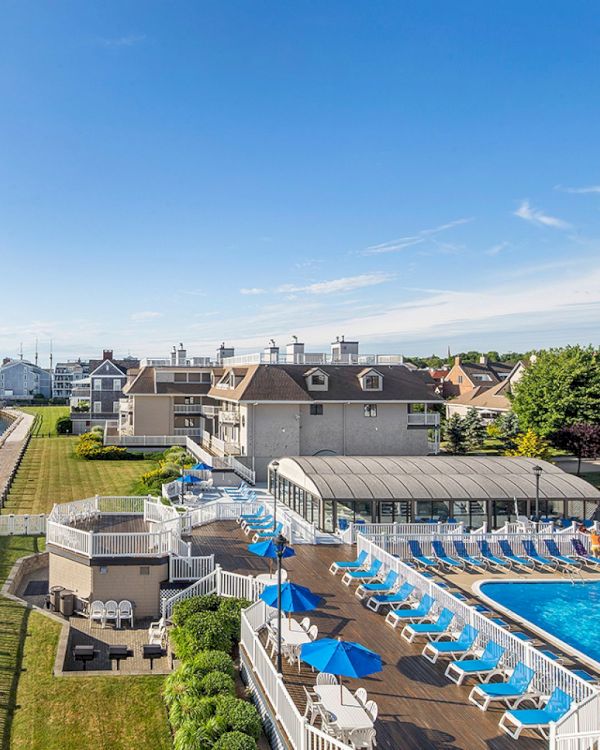 A coastal resort with a pool and blue umbrellas overlooks the water, surrounded by greenery and adjacent buildings, under a clear sky.