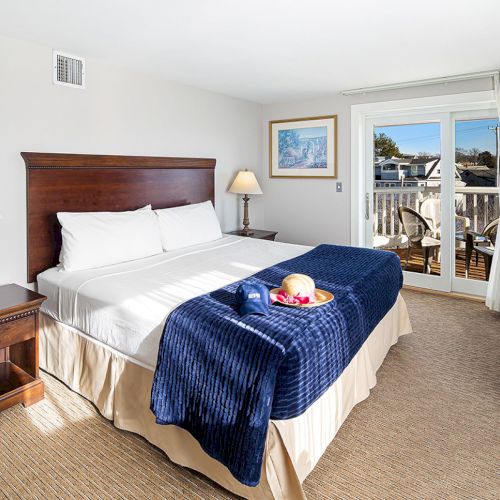 A hotel room with a king bed, blue blanket, bedside tables, lamps, and a balcony view. A tray with fruit and wine on the bed completes the scene.