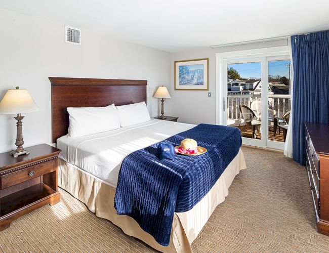 A hotel room with a king bed, blue blanket, bedside tables, lamps, and a balcony view. A tray with fruit and wine on the bed completes the scene.