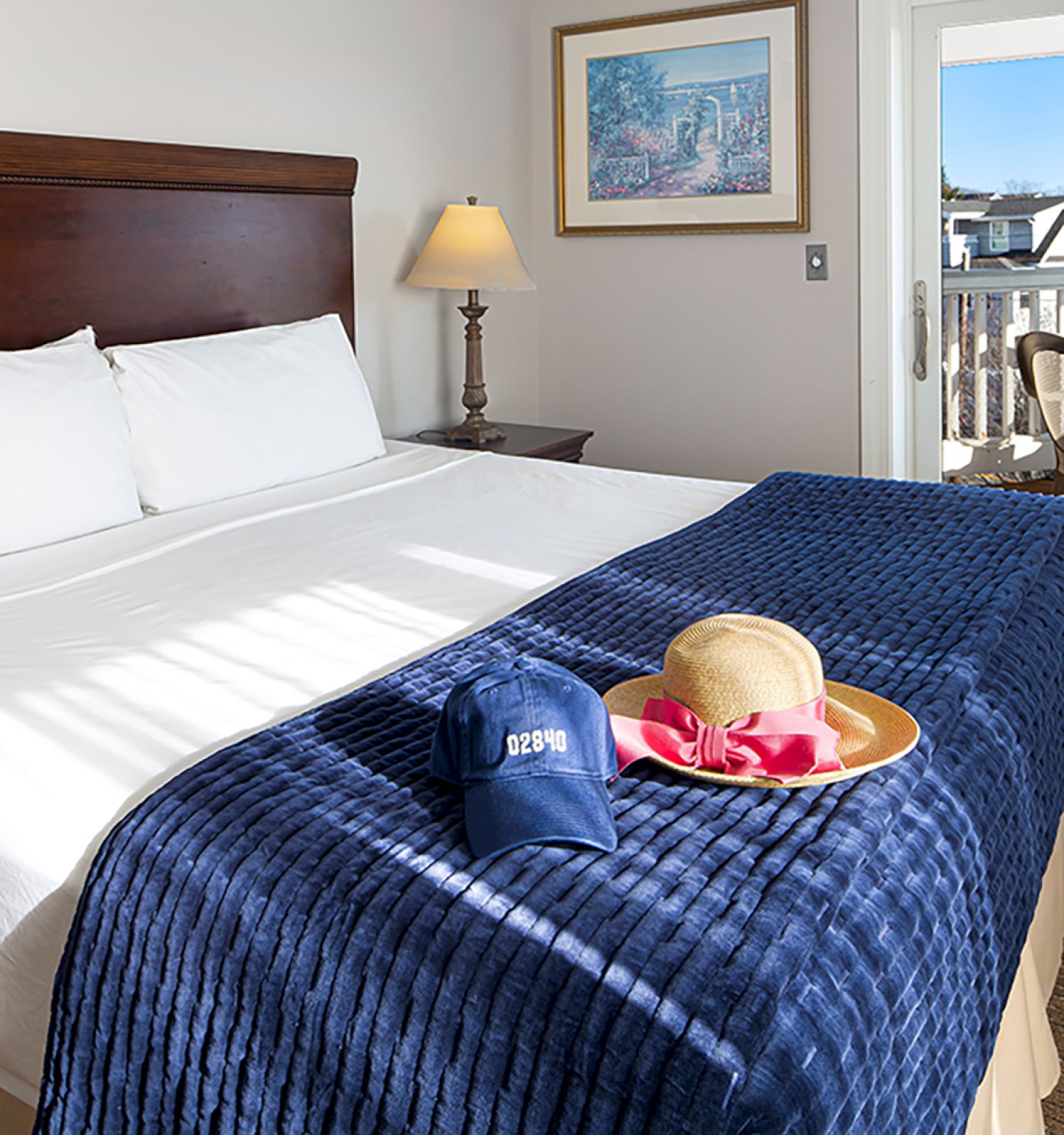 A neatly made bed with a blue blanket, a hat, and snacks on a tray. There's a lamp, painting, and a view through the window in the background.