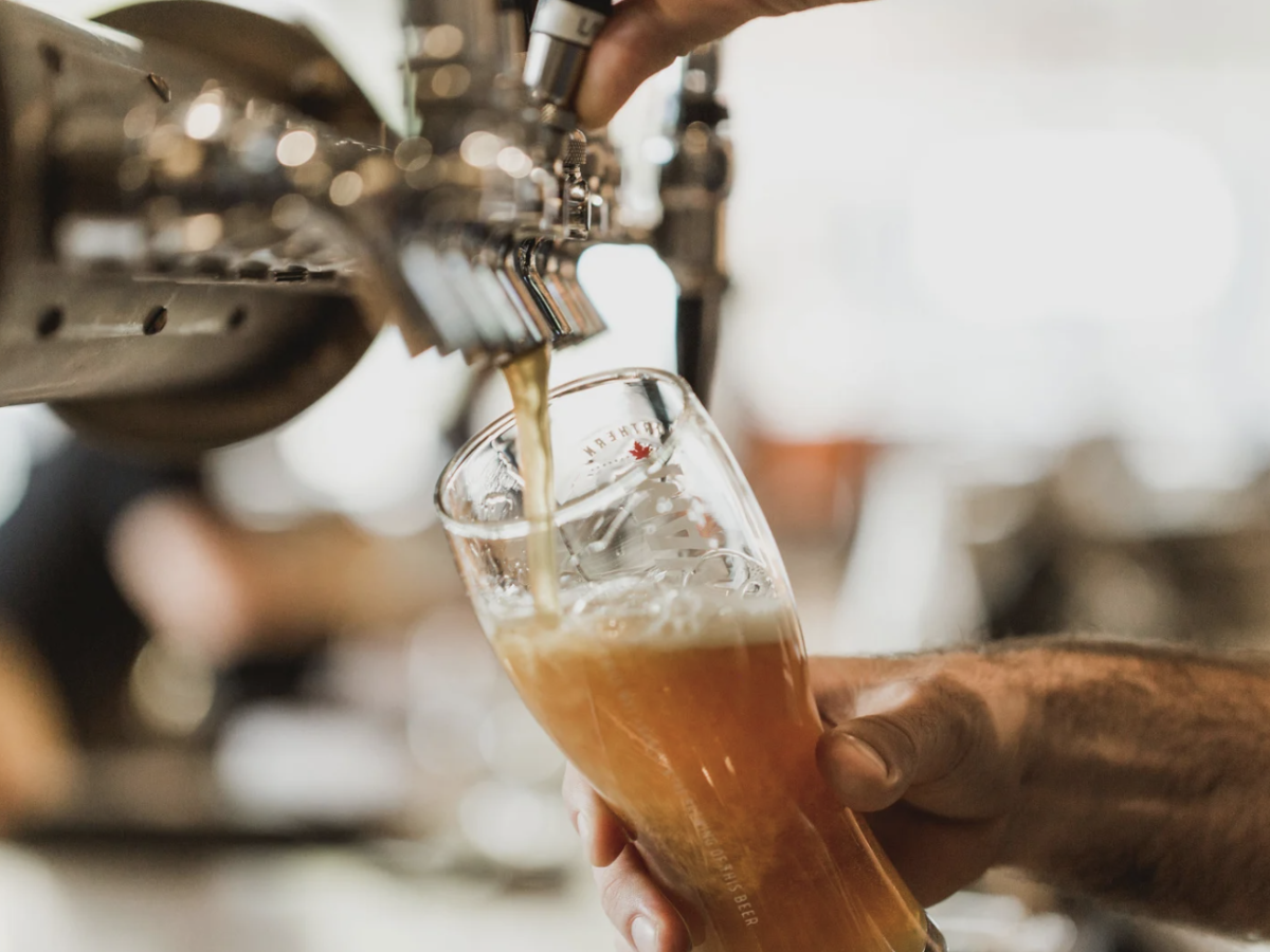 A hand pours a draft beer into a tilted glass from a tap, creating a foamy top layer in a bar or pub setting.