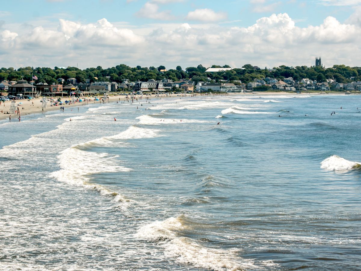 A sandy beach with people, surf waves, and nearby buildings surrounded by greenery under a partly cloudy sky.