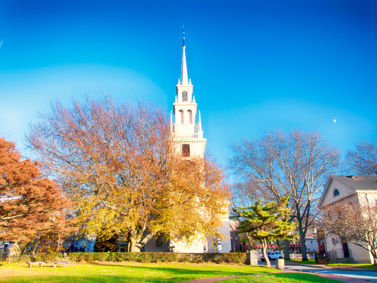 A church with a tall steeple stands amid trees in autumn, under a clear blue sky, with a grassy area in the foreground.