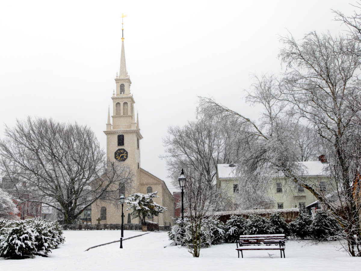 A snowy landscape featuring a church with a clock tower, surrounded by leafless trees and buildings.