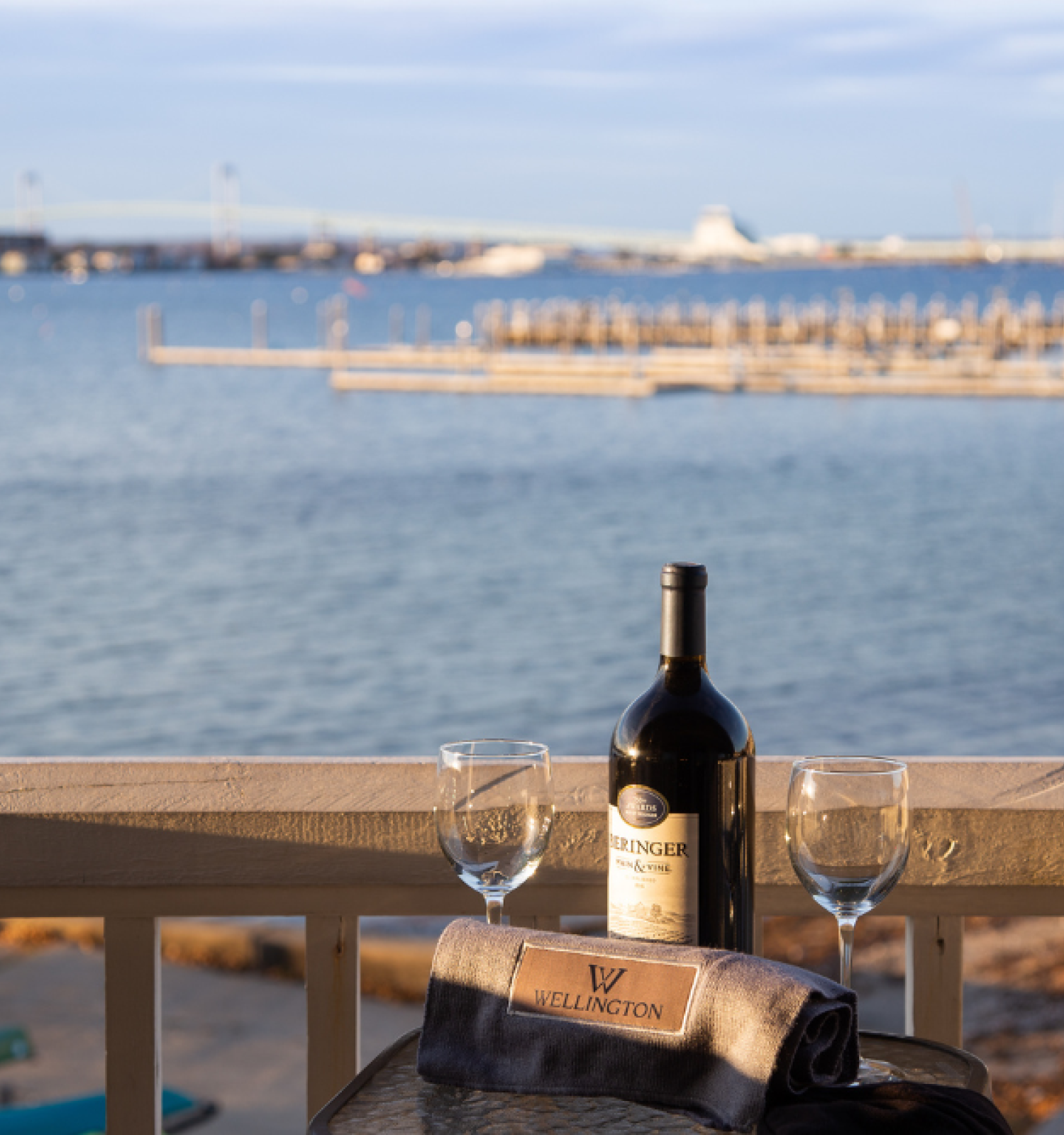 A bottle of wine and two glasses on a table overlook a scenic waterfront with a marina in the distance, under a clear blue sky.